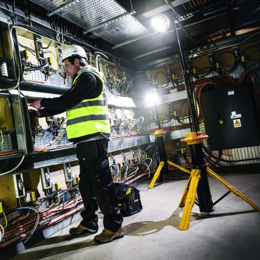 Worker wearing safety clothing in am electrics room with 2 Defender light cannons in the background illuminating the room